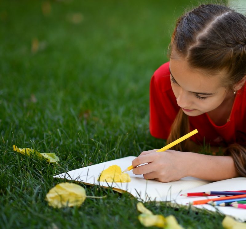 A teenage girl lies on the grass and draws with colorful pencils.